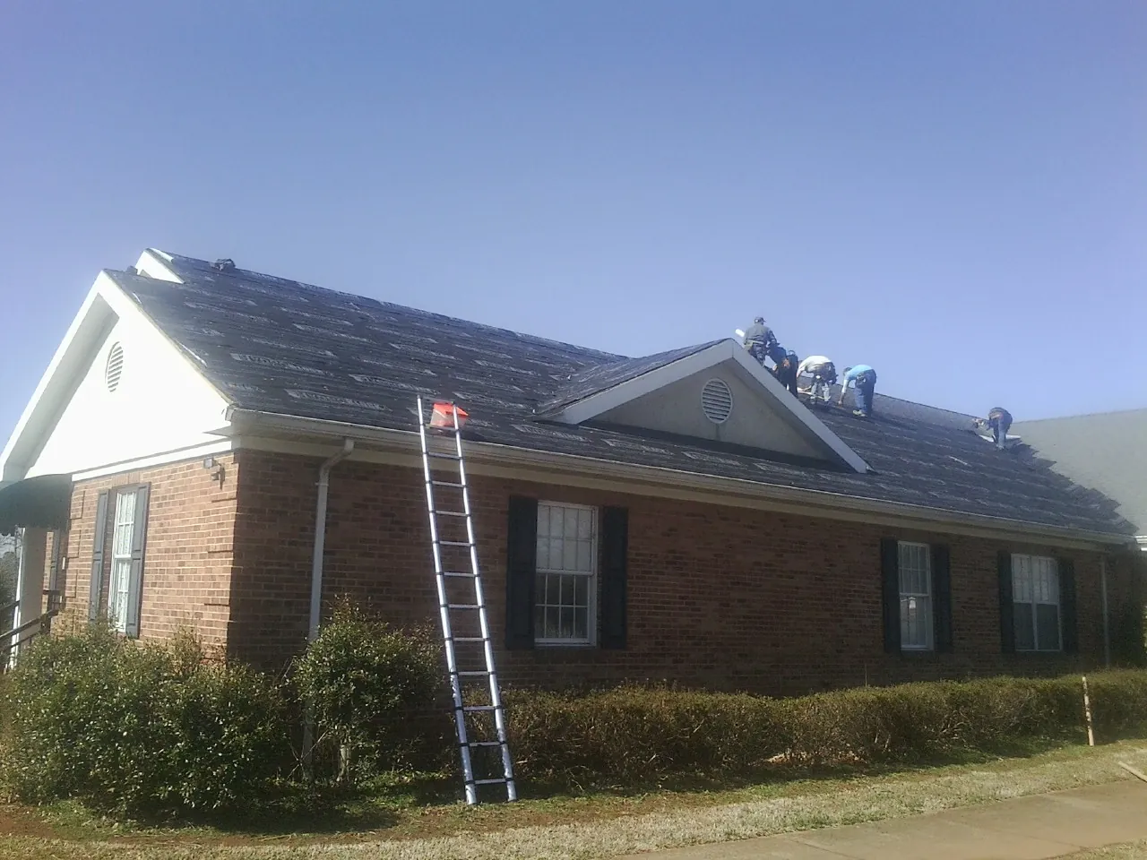 Crew tearing shingles off a brick ranch roof with a ladder set — Emergency Roof Repair work by our crew in Le Claire