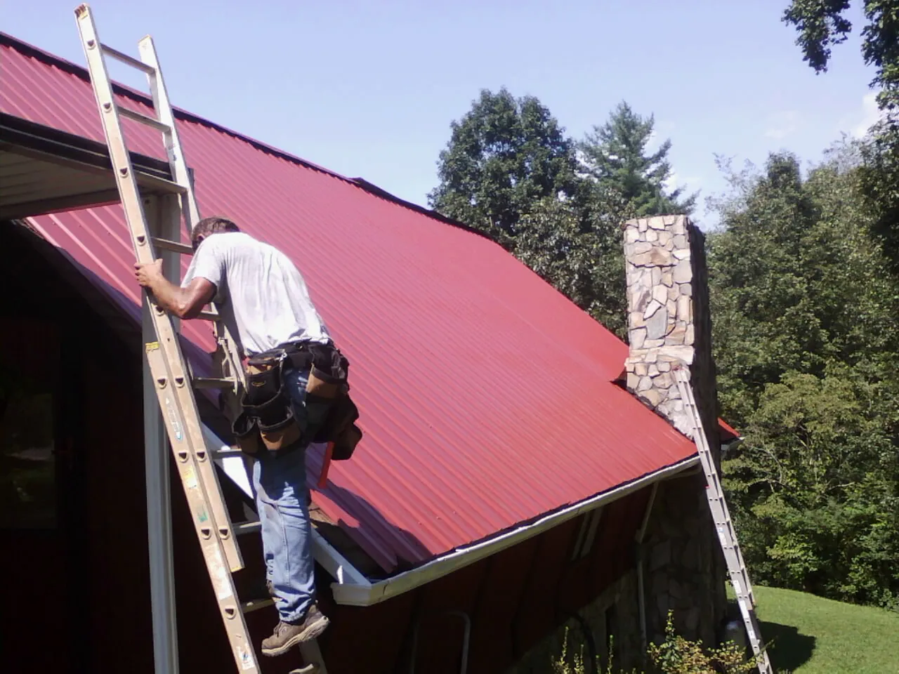 Roofer on a ladder beside a red metal roof and stone chimney — Soffit & Fascia Repair by Roofing Brothers in Le Claire